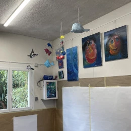 Espace atelier enfants - <p>L'espace enfants est lumineux et coloré, avec des mobiles d'animaux marins suspendus au plafond. Une table basse, "la table des couleurs", invite à la création avec ses pots de peinture. Des œuvres d'art enfantines décorent les murs. Une bâche bleue protège le sol.</p> - Nathalie Vazquez
